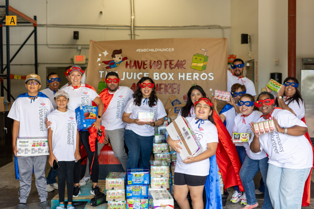 A group of volunteers from Bank of America wearing superhero capes and masks pose together in front of a banner that reads 'Have no fear, the Juice Box Heroes are here!' in the Kids' Meals warehouse.