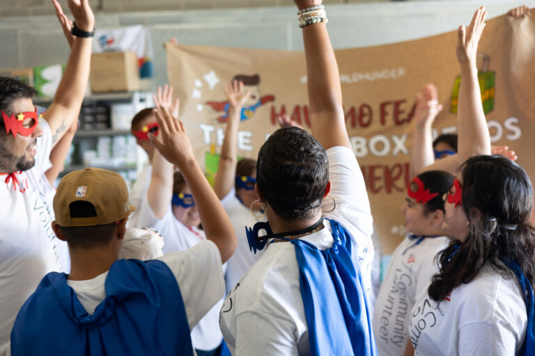 Bank of America Volunteers in superhero capes and masks raise their hands in a team cheer in front of a Juice Box Heroes banner